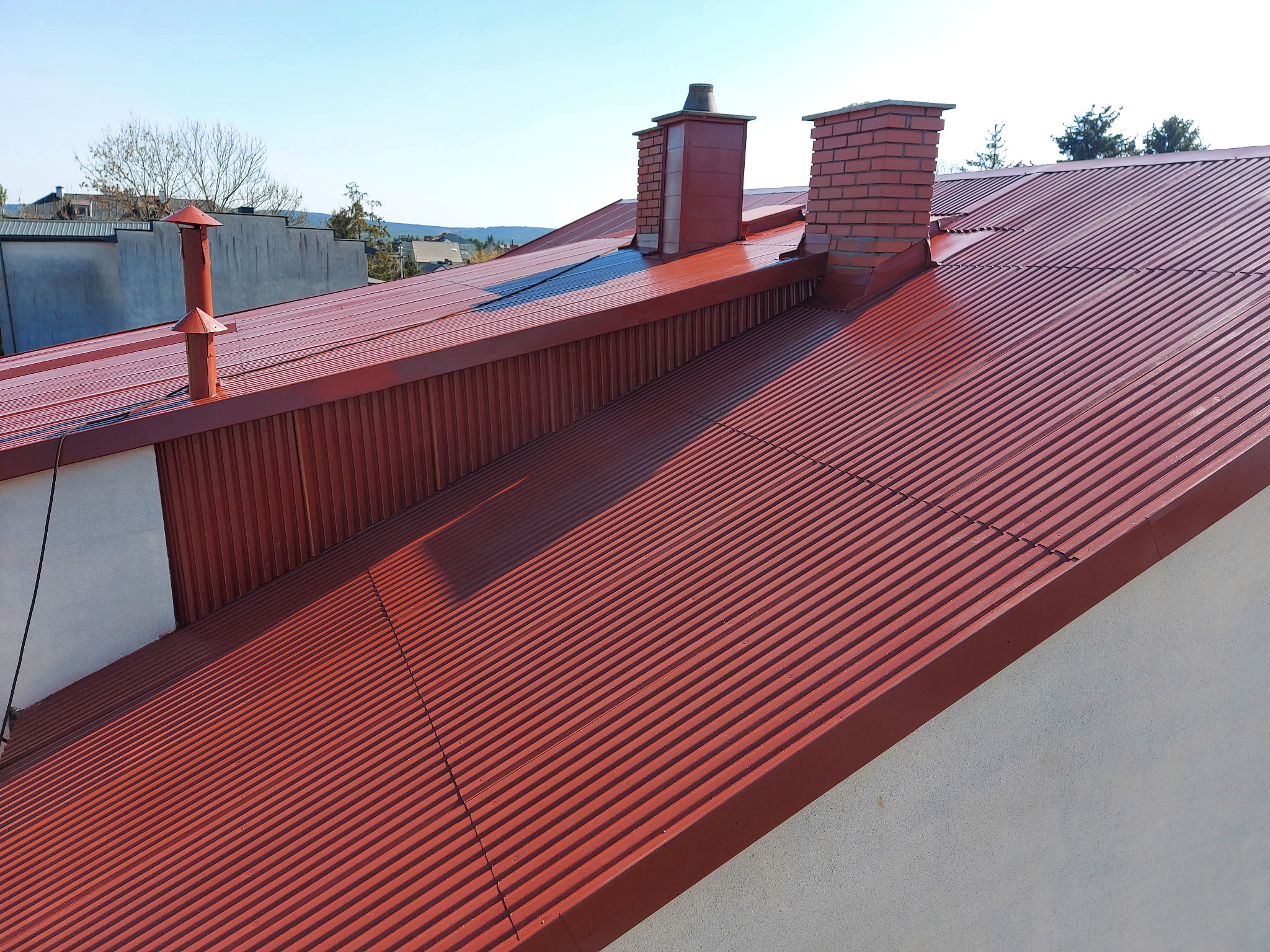 Czerwony, blaszany dach z dwoma ceglanymi kominami i jednym metalowym odpowietrznikiem na tle błękitnego nieba.