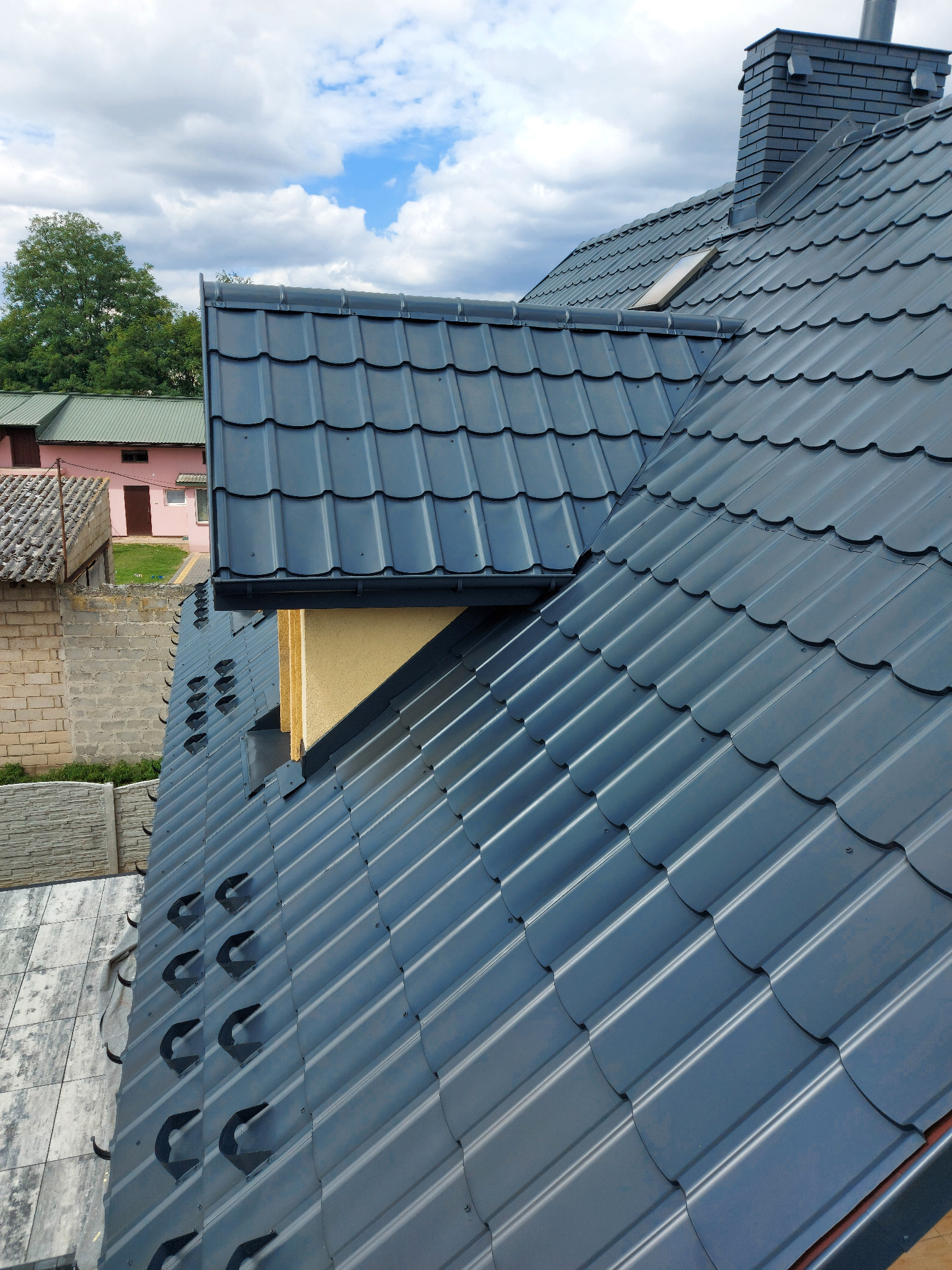 Nowy, grafitowy dach z blachodachówki modułowej z kominem w kolorze dachu i oknem dachowym, widok z góry na tle błękitnego nieba z chmurami.