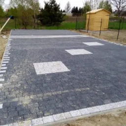 Ułożona, szara kostka brukowa z białymi elementami dekoracyjnymi na podwórku z drewnianą szopą w tle, widok z góry.