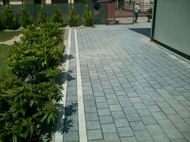 Szara kostka brukowa ułożona wzdłuż granicy działki, z widocznymi roślinami i fragmentem ogrodzenia w tle, tworząca estetyczny podjazd.