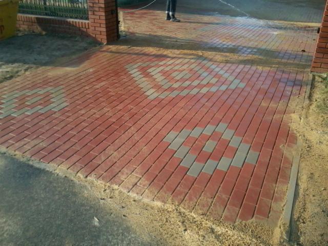 Czerwona kostka brukowa ułożona we wzór z szarymi elementami, tworząca podjazd. Widoczny fragment muru z cegły i piasek przy krawędziach.