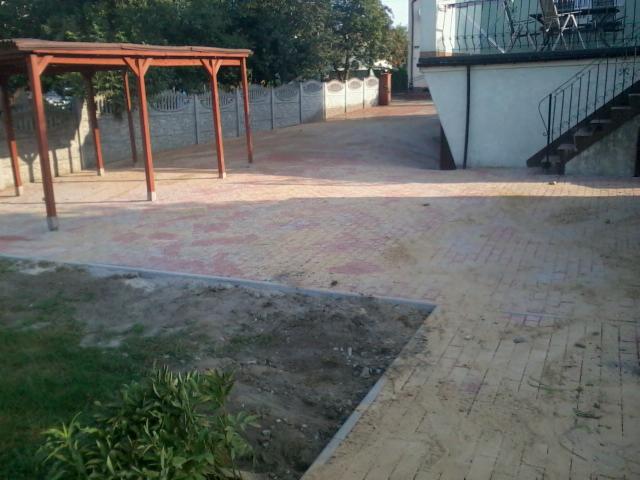 Ukończona nawierzchnia z kostki brukowej w odcieniach czerwieni, z drewnianą pergolą w tle, obok budynku z metalowymi schodami i szarą, betonową ścianą.