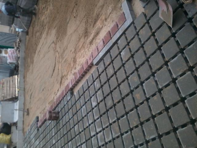 Częściowo ułożona, kwadratowa, szara kostka brukowa z czerwoną obwódką na podsypce piaskowej. Widoczna łopata i paleta z kostką.