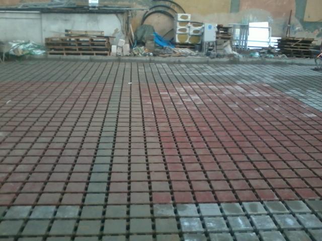 Częściowo ułożona, ażurowa kostka brukowa w odcieniach czerwieni i szarości na placu budowy. Widoczne palety i materiały budowlane w tle.