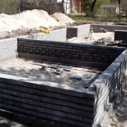 Wznoszone ściany fundamentowe z bloczków betonowych na piaszczystym podłożu, widoczny wykop i poziomica laserowa, w tle hałdy piasku.