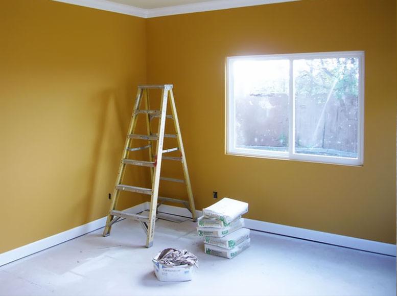 Żółta ściana świeżo po malowaniu, widoczna drabina, worki z materiałem budowlanym i pojemnik z papierem zabezpieczającym podłogę.