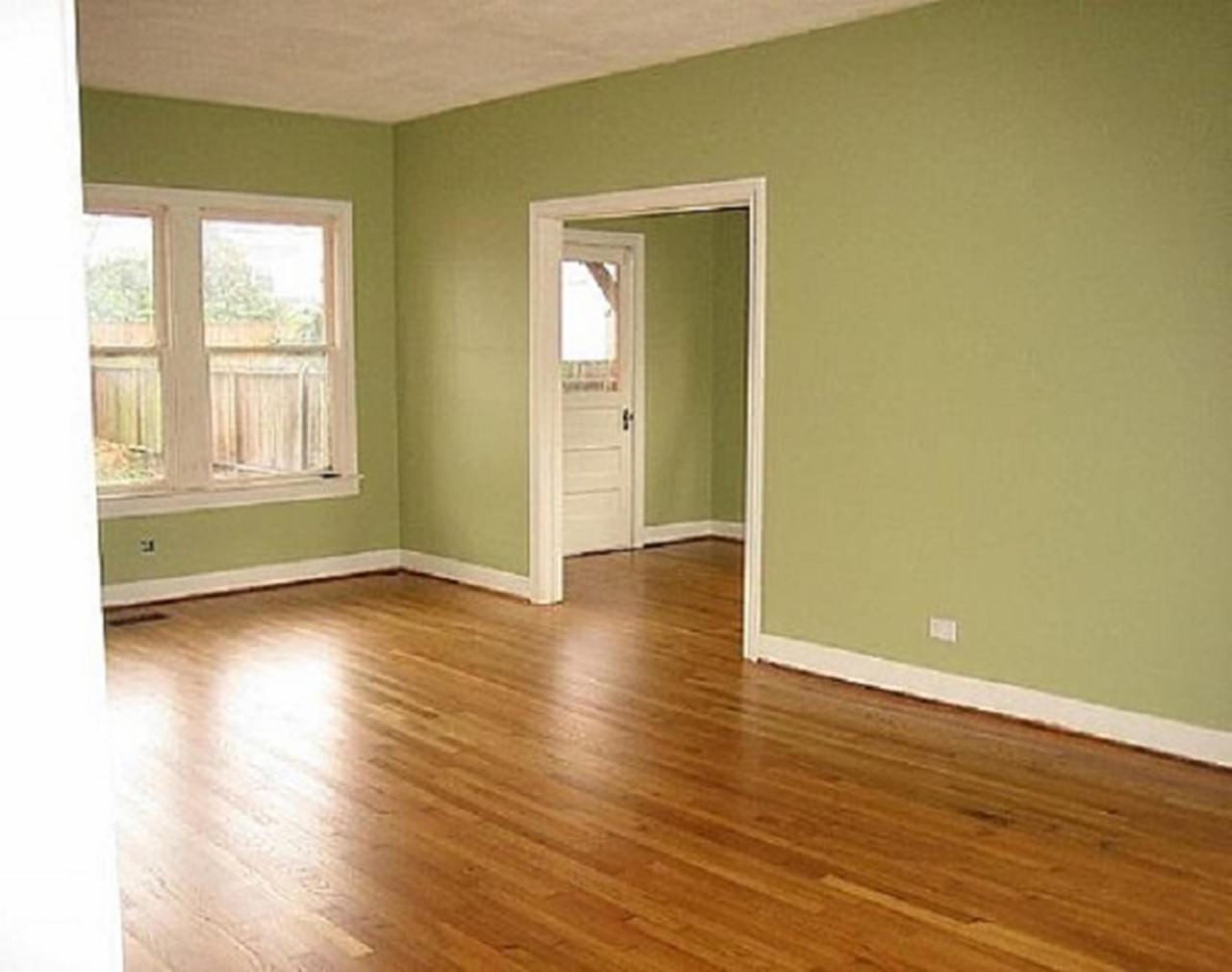 Wnętrze pokoju z drewnianą podłogą i ścianami pomalowanymi na pistacjowy kolor; widoczne okno z białymi ramami i drzwi do innego pomieszczenia. Listwy przypodłogowe są białe, a na ścianie widoczny...