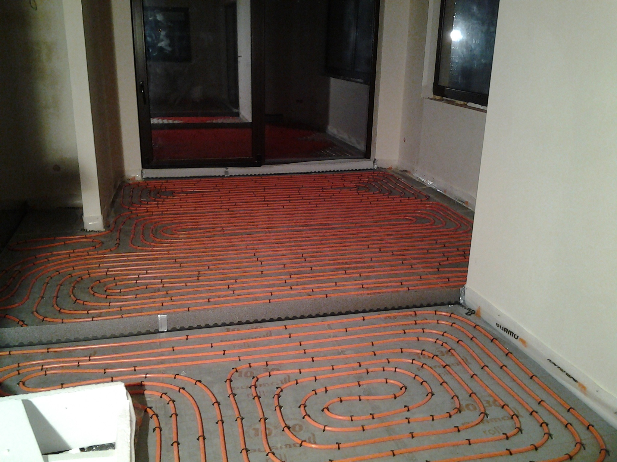 Pomarańczowe rury ogrzewania podłogowego ułożone na szarych płytach izolacyjnych w jasnym pomieszczeniu z dużym oknem balkonowym, widoczne czarne uchwyty mocujące rury do podłoża.