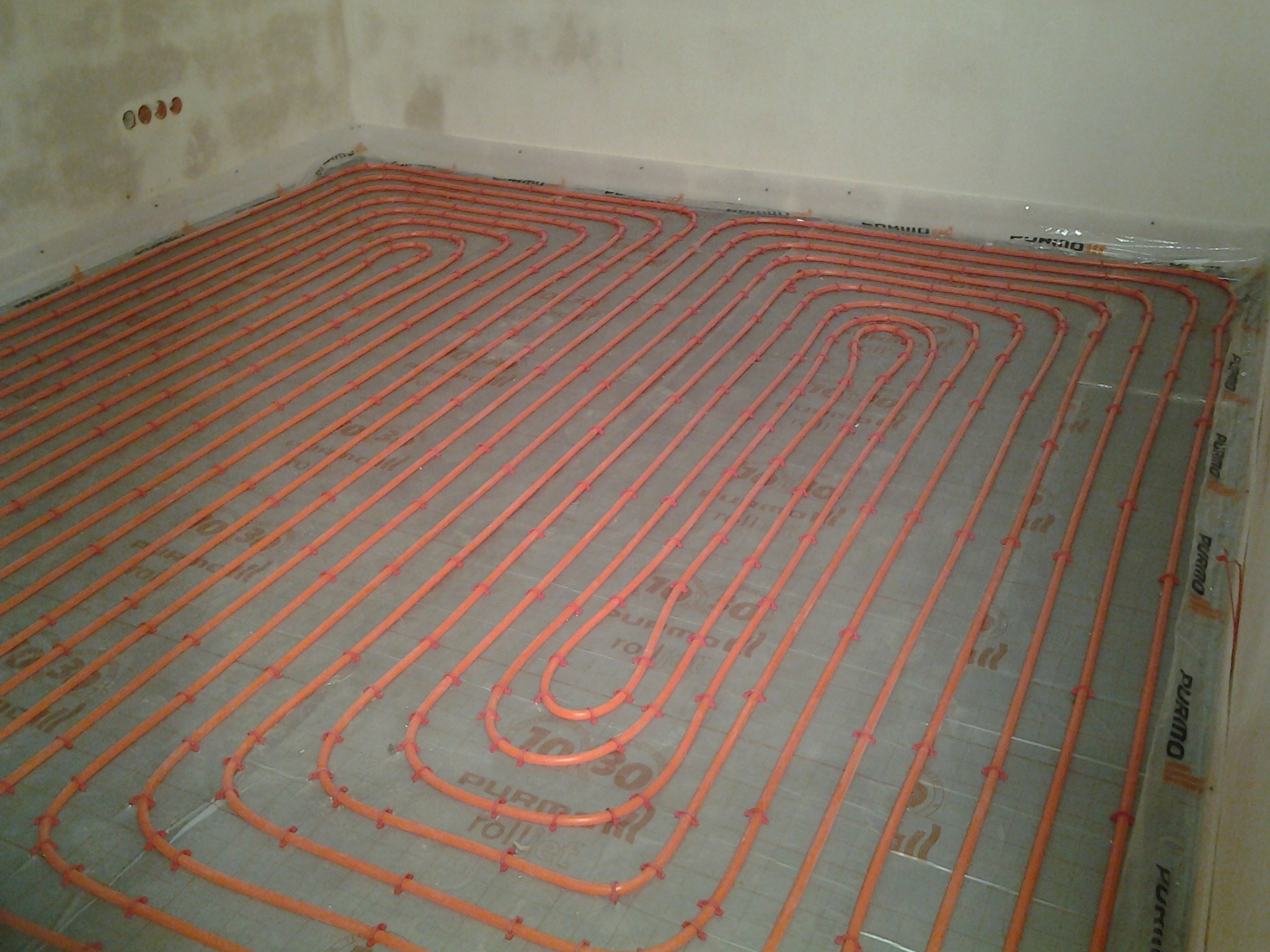 Pomarańczowe rury ogrzewania podłogowego ułożone w pętle na szarej izolacji z nadrukiem, widoczne mocowania rur, surowy stan wykończenia ścian.