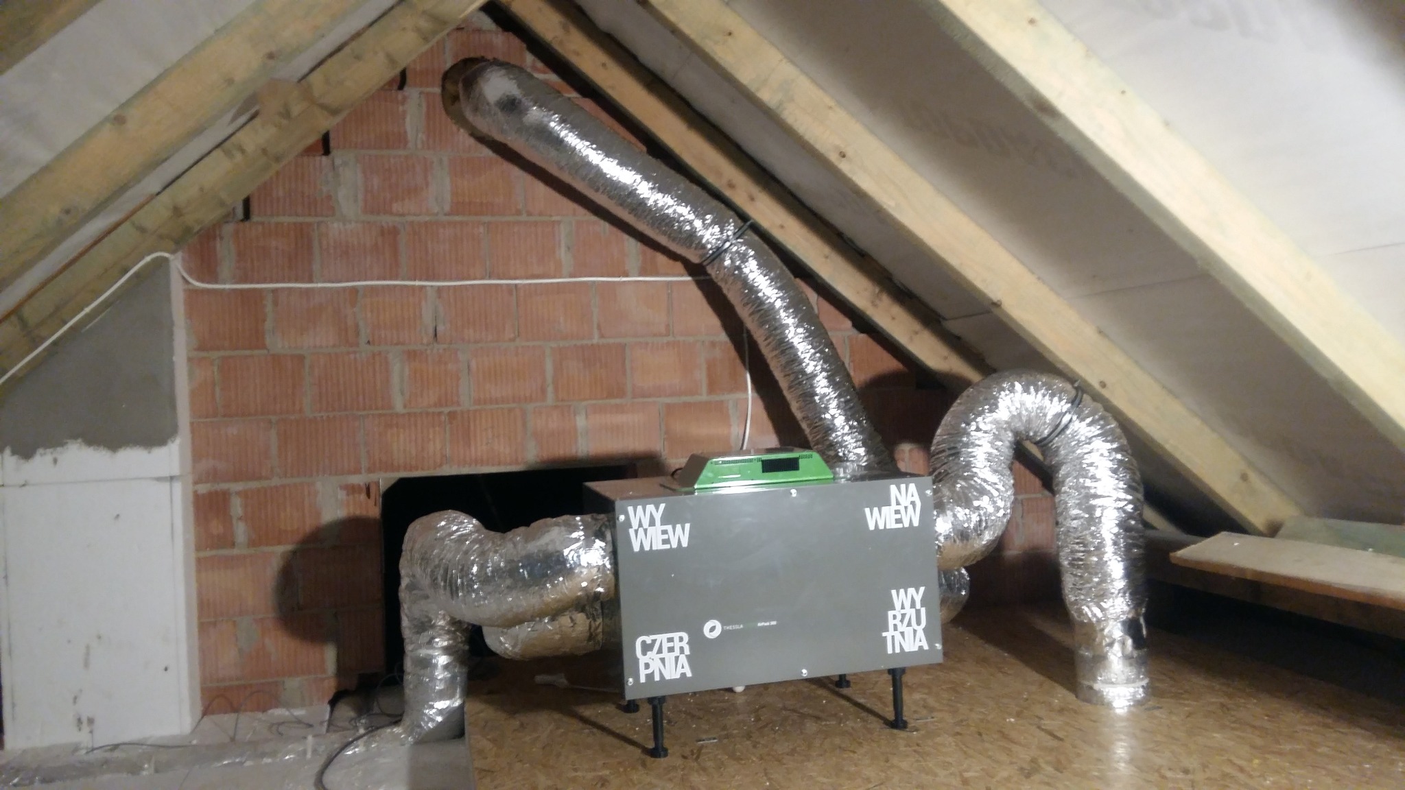 Szara centrala wentylacyjna z podłączonymi srebrnymi przewodami wentylacyjnymi w przestrzeni poddasza, widoczne drewniane elementy konstrukcji dachu i fragment ściany z cegły.