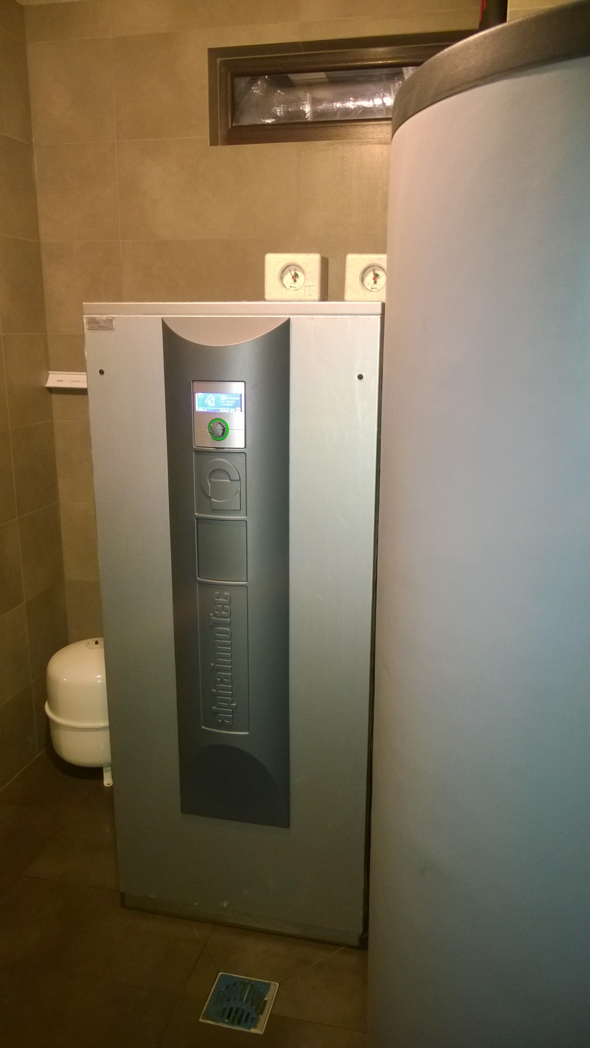 Szara pompa ciepła Alphainnotec w pomieszczeniu z beżowymi płytkami, widoczny zbiornik wyrównawczy i dwa manometry nad urządzeniem.