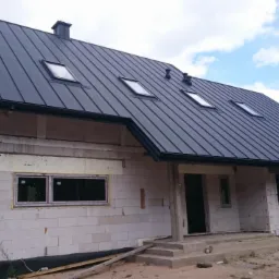 Dom w trakcie budowy z ciemnym, metalowym dachem z oknami dachowymi. Ściany z białego bloczka częściowo ocieplone. Widoczne okna i drzwi.