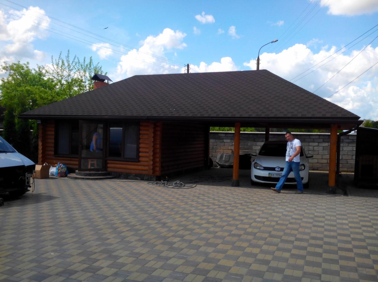 Dom z bali z ciemnobrązowym dachem bitumicznym i zadaszonym miejscem parkingowym. Mężczyzna przechodzi obok białego samochodu. Kostka brukowa na podjeździe.