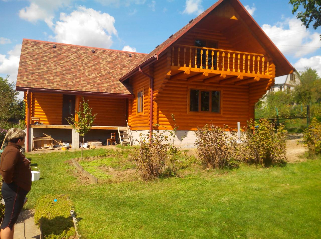Dwukondygnacyjny dom z bali z balkonem, wykończony czerwoną dachówką bitumiczną. Widoczna kobieta w ogrodzie. Błękitne niebo z chmurami w tle.