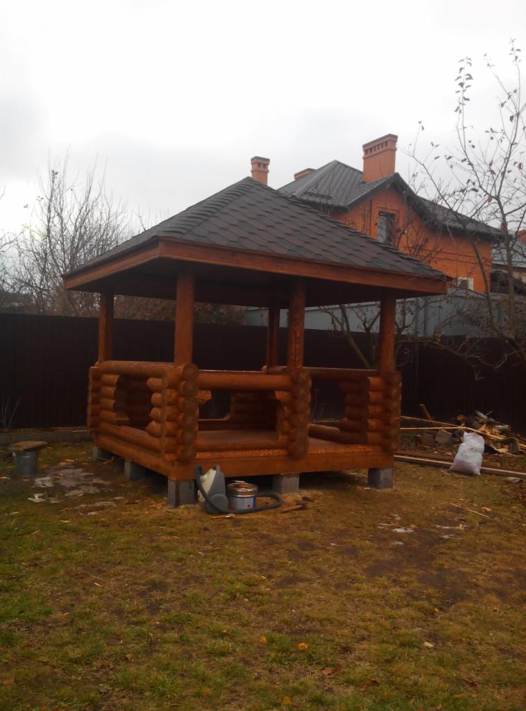 Drewniana altana ogrodowa z bali w Krakowie, z ciemnym dachem bitumicznym, na tle domu z czerwonej cegły i brązowego ogrodzenia. Widoczne narzędzia i resztki materiałów.