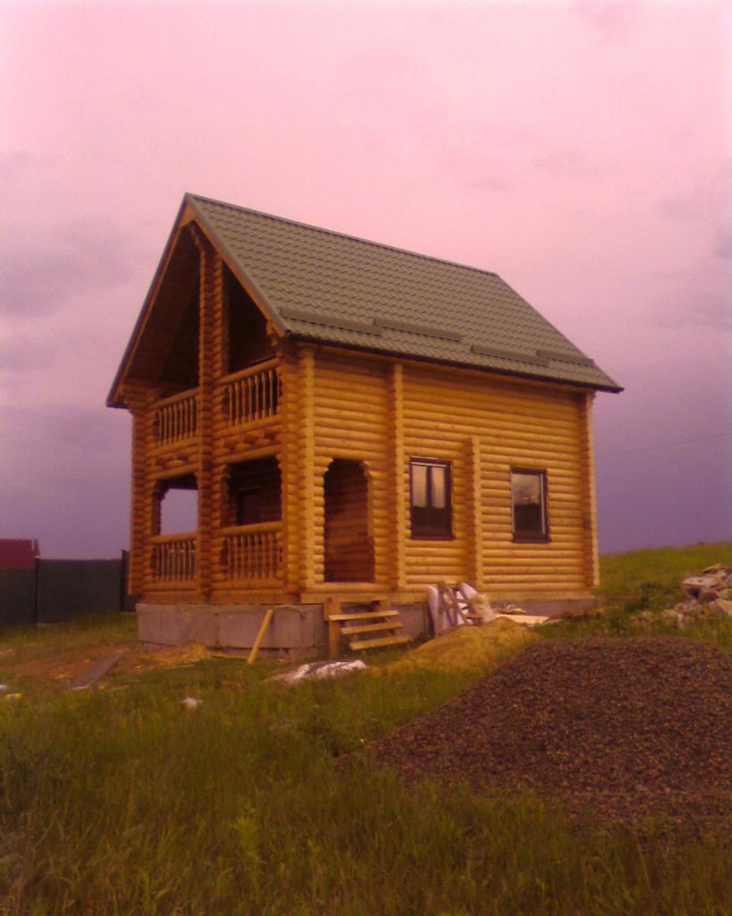 Dwukondygnacyjny, drewniany dom z bali z zielonym dachem na wzgórzu, pod różowym niebem. Widoczne schody wejściowe i sterta żwiru.