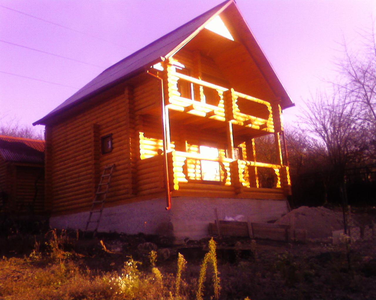 Dwukondygnacyjny dom z bali, z widoczną konstrukcją balkonu i drabiną opartą o ścianę. Budynek na wzniesieniu, w tle drzewa bez liści, fioletowe niebo.