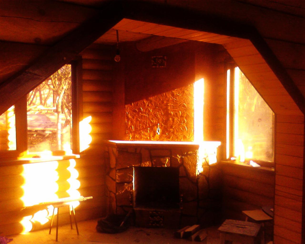 Wnętrze domu z bali z kominkiem w kamiennej obudowie. Widoczne okna wpuszczające silne promienie słoneczne. Drewniane ściany i sufit. Narożny układ pomieszczenia.