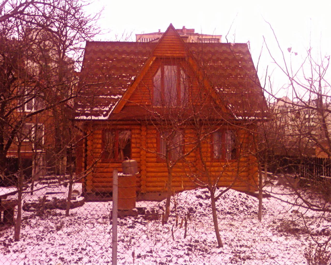 Zimowy krajobraz z drewnianym domem z bali o spadzistym dachu z brązowej dachówki, na pierwszym planie ośnieżone drzewa i metalowa beczka.