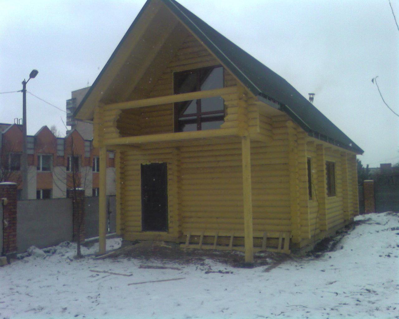 Żółty dom z bali z ciemnym dachem na tle zaśnieżonego krajobrazu miejskiego. Widoczny balkon i okno z geometrycznym podziałem.