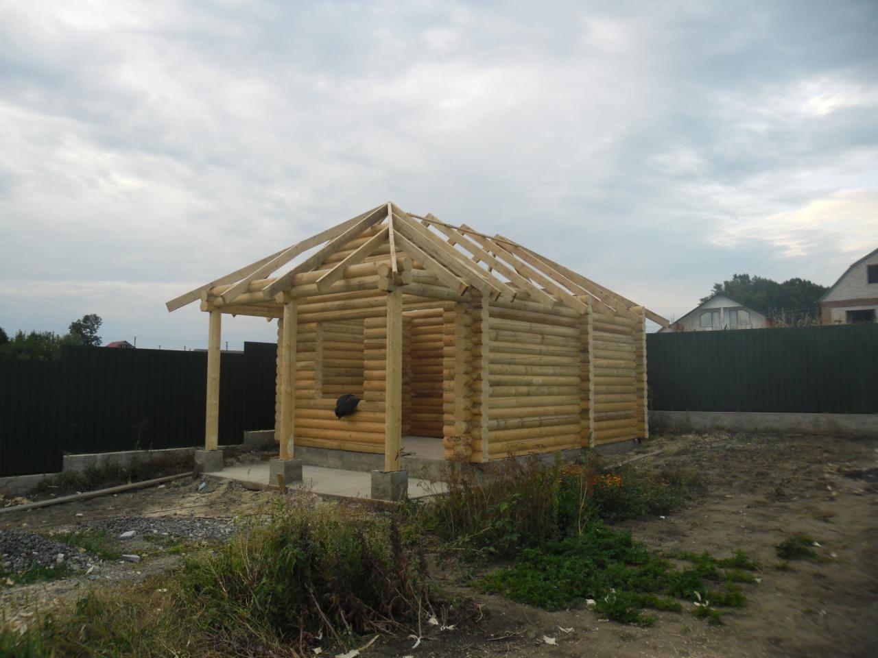 Surowy stan domu z bali w trakcie budowy, widoczne ściany i więźba dachowa na tle pochmurnego nieba i zielonego ogrodzenia.