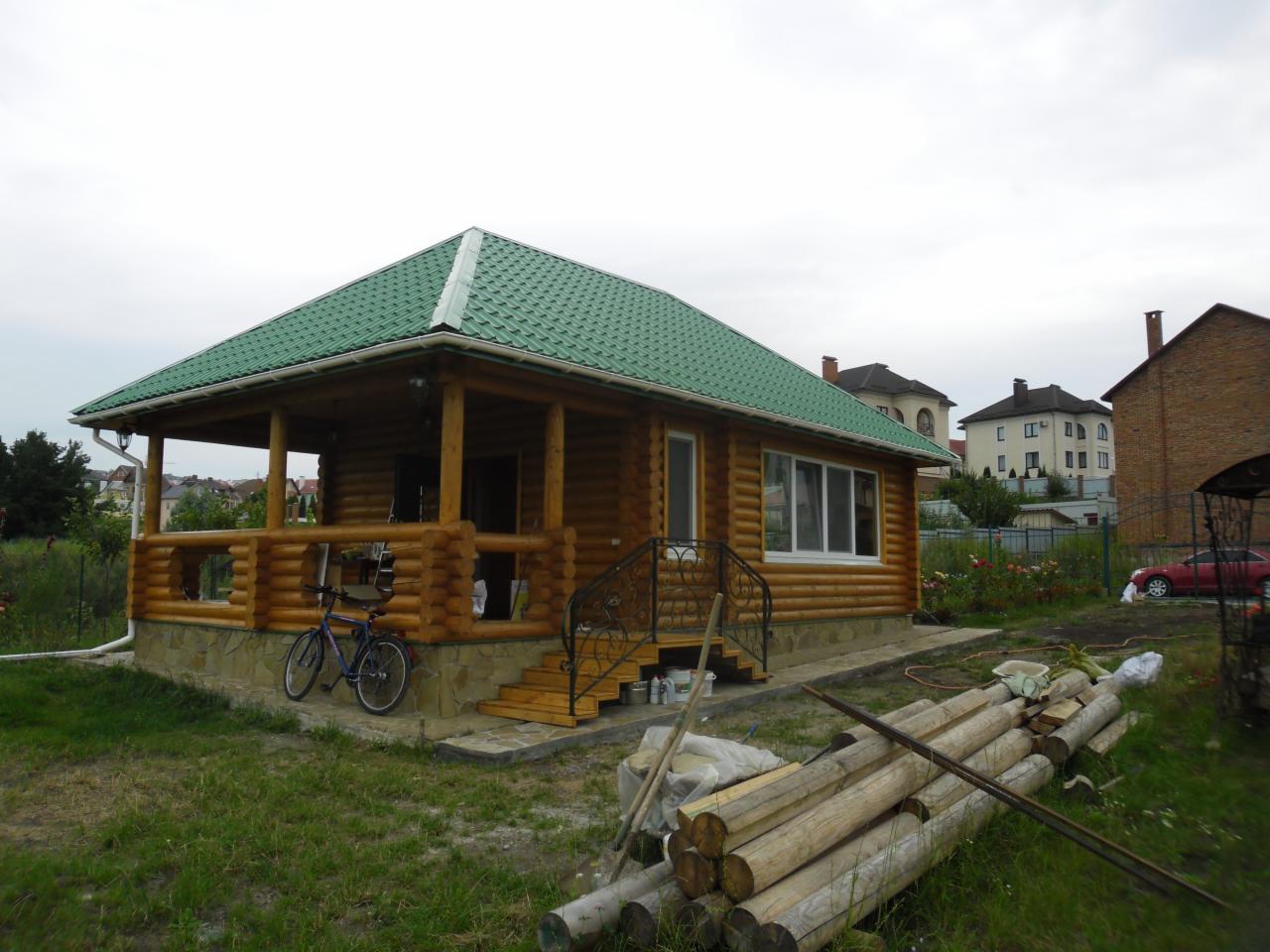 Drewniany dom z bali z zielonym dachem, widoczne schody z kutą balustradą i rower oparty o werandę. Na trawie leżą przygotowane bale.