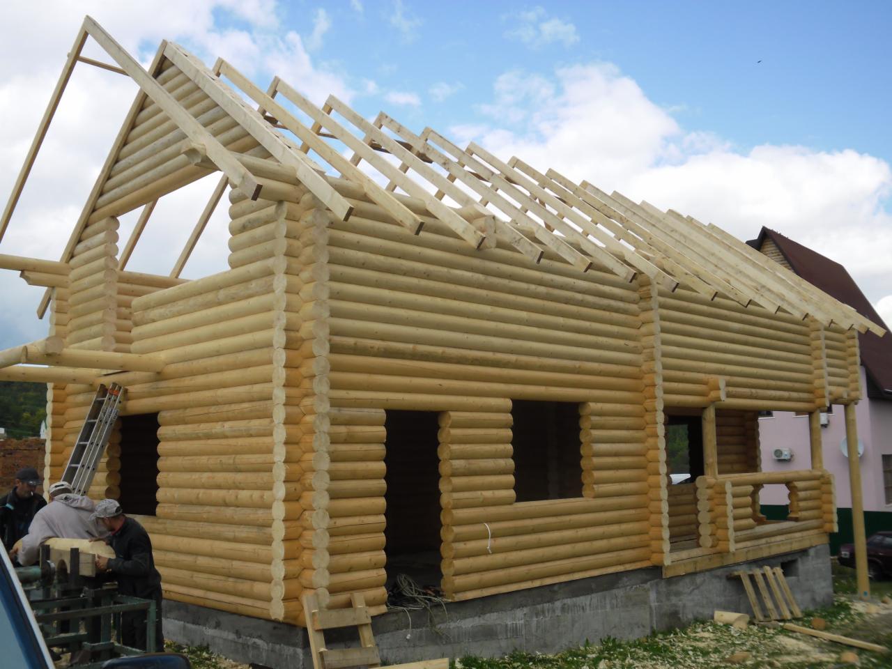 Surowy dom z bali w trakcie budowy, widoczne ściany z bali, konstrukcja dachu i fundament. Na pierwszym planie pracownicy z drewnianymi elementami.