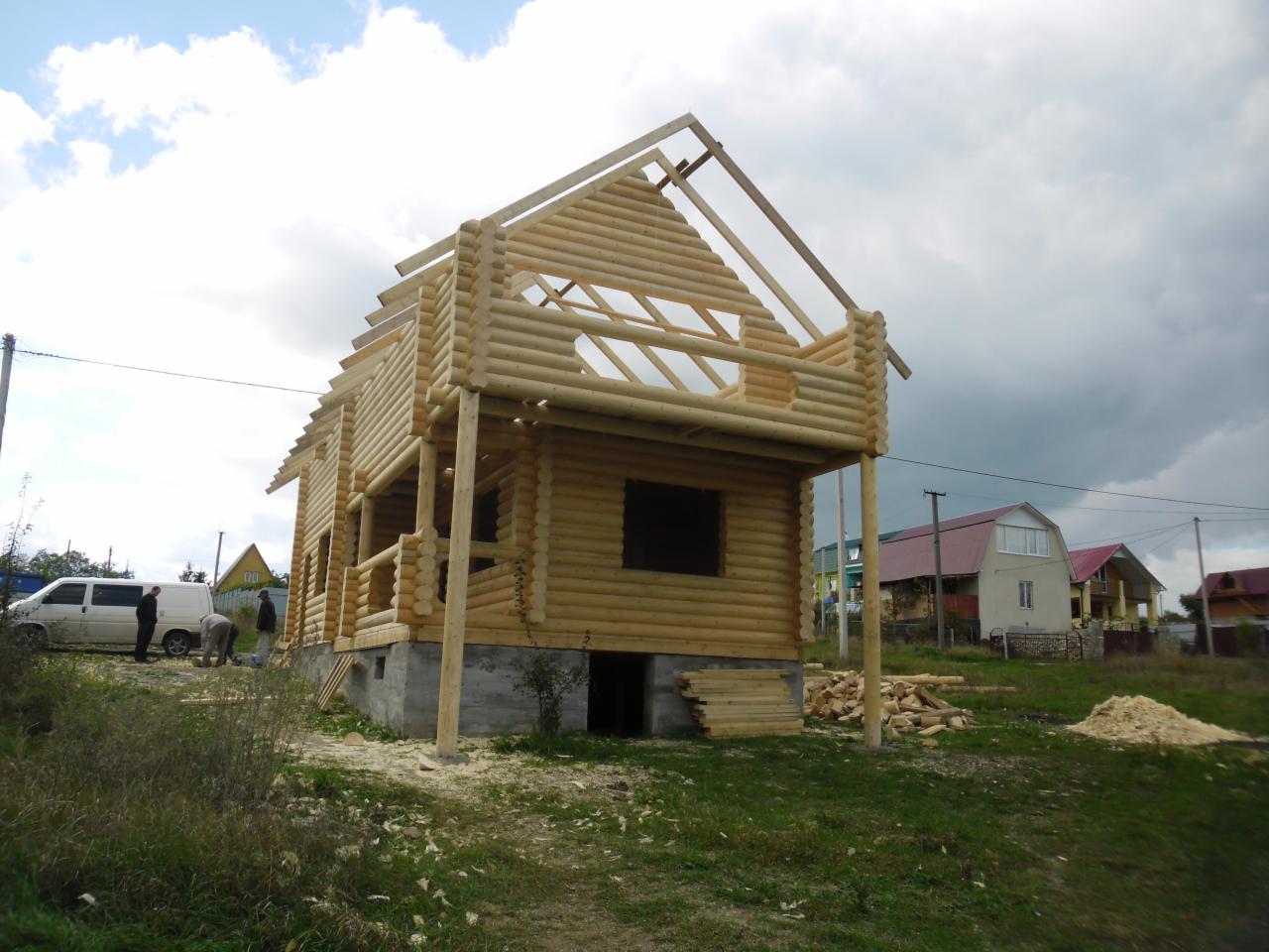 Surowy dom z bali w trakcie budowy, widoczne drewniane elementy konstrukcyjne, fundament i pracownicy. Tło stanowią inne domy i pochmurne niebo.