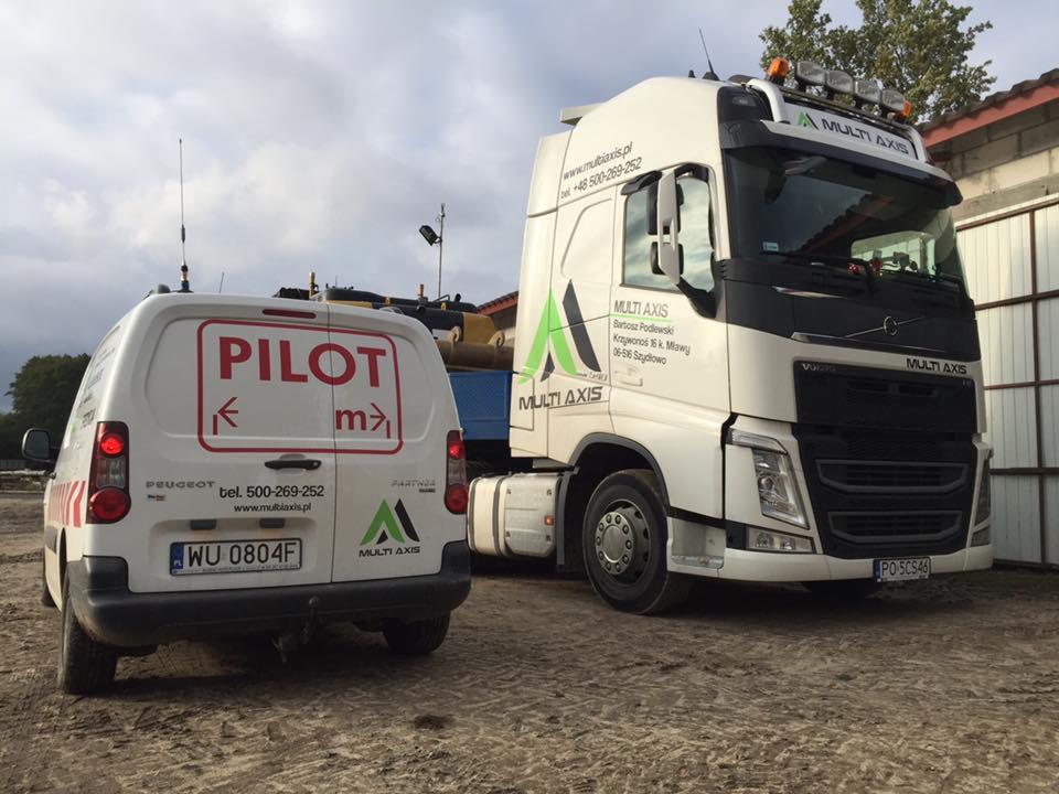 Ciężarówka Volvo z naczepą oraz samochód pilotujący Peugeot Partner z napisem PILOT, oba pojazdy z logo firmy Multi Axis, stojące na placu.