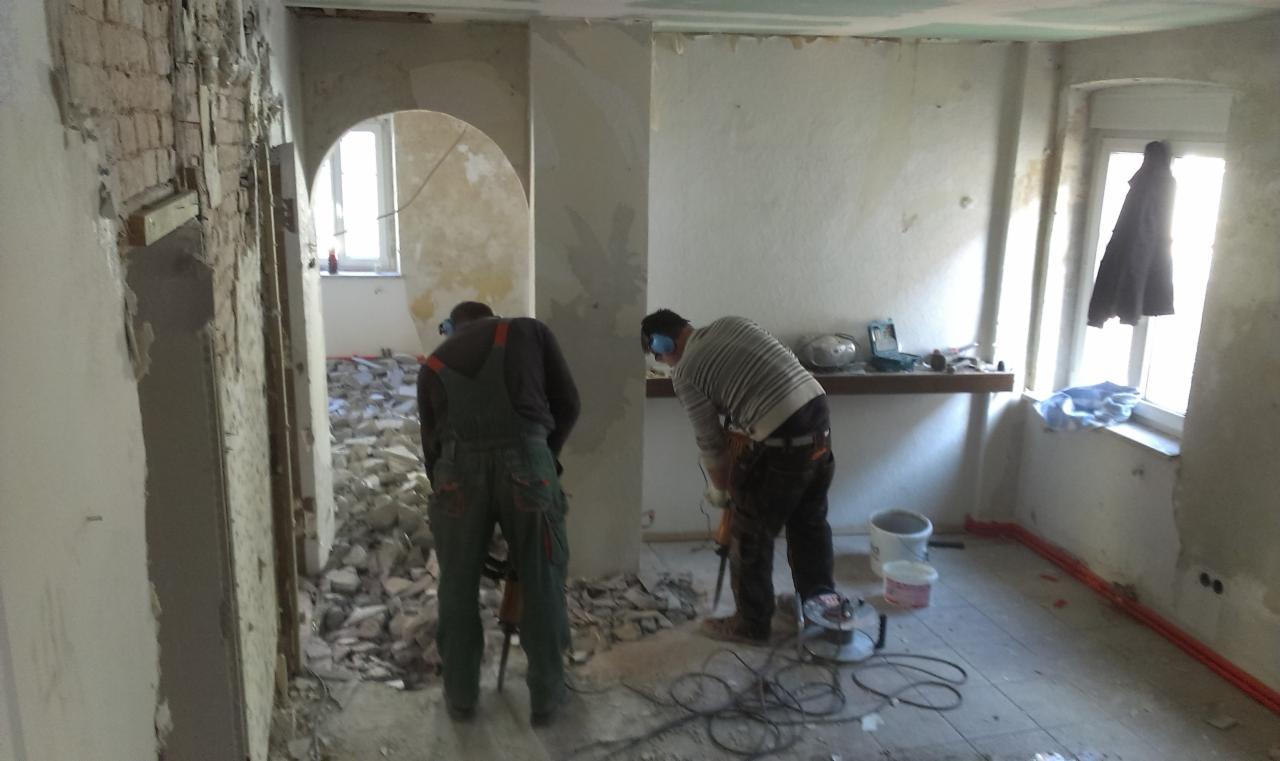 Dwóch robotników w trakcie skuwania tynku ze ścian w remontowanym pomieszczeniu z widocznymi narzędziami i gruzem. Prace remontowe w starym budynku.