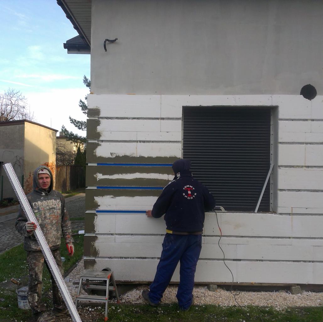 Ocieplanie domu: dwóch mężczyzn montuje izolację z białych bloków, używając poziomicy. Widoczne okno z roletą i elementy budynku w trakcie prac.