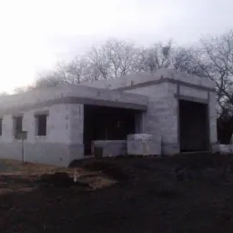 Surowy stan budynku z betonu komórkowego, widoczne niezabezpieczone otwory okienne i drzwiowe, hałda piasku z boku i worki z materiałem budowlanym przed budynkiem, w tle drzewa bez liści.