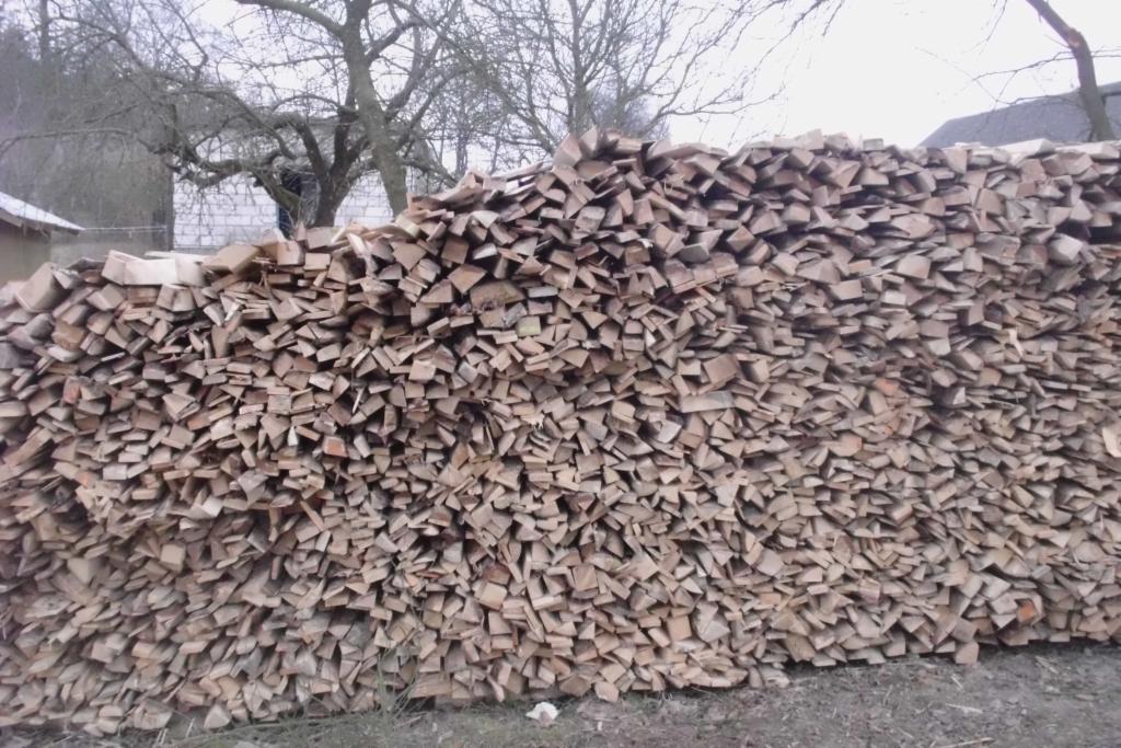 Równomiernie ułożony stos pociętego drewna kominkowego na zewnątrz, z widocznymi śladami kory, na tle drzew bez liści i zabudowań.