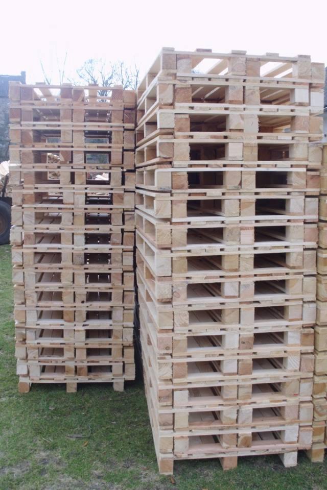 Dwie stosy jasnych, drewnianych palet na trawie, widoczne elementy pojazdu w tle. Palety ułożone równo, z wyraźną strukturą drewna i przestrzeniami między deskami.