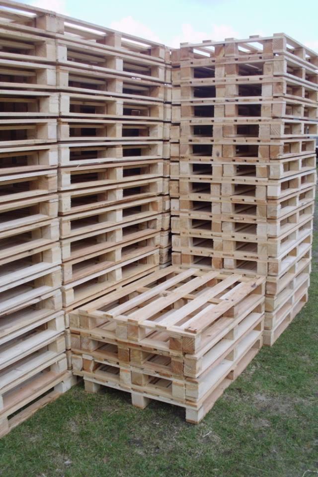 Stos drewnianych palet euro na trawie, widok z dołu. Palety ułożone w rzędach i kolumnach, tworzą regularny wzór. Naturalne oświetlenie, widoczna struktura drewna.