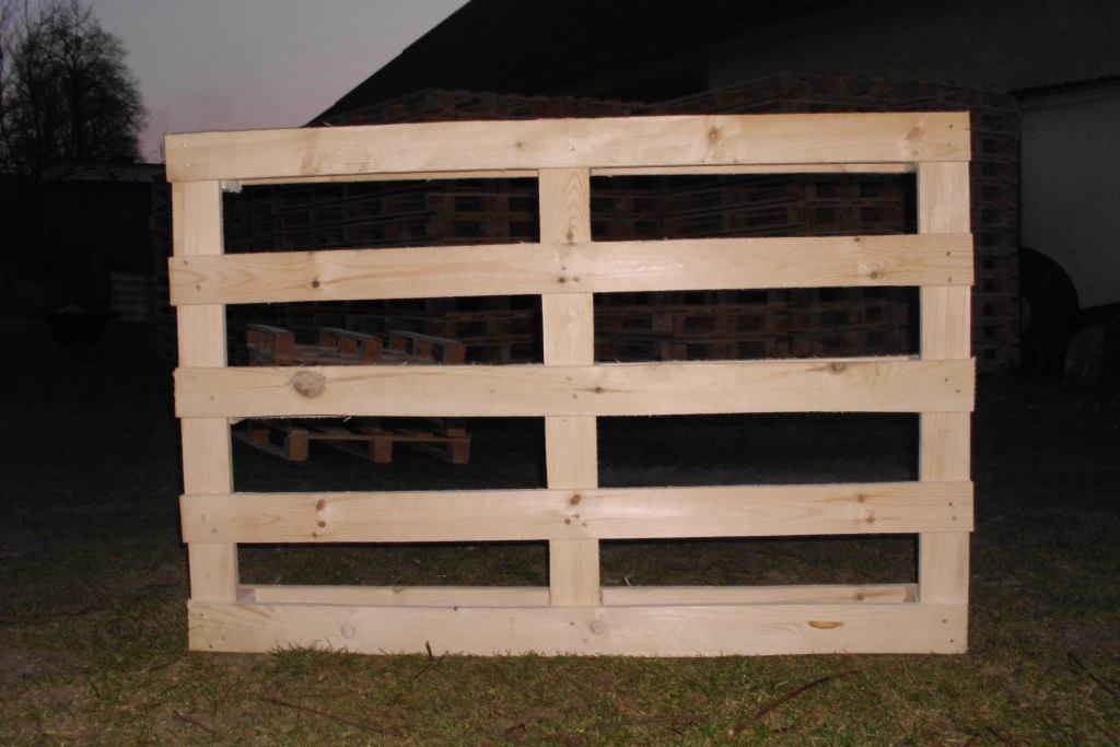 Surowa, jasna paleta drewniana na trawie, widok z przodu. W tle stos palet i fragment budynku. Proste, solidne wykonanie.