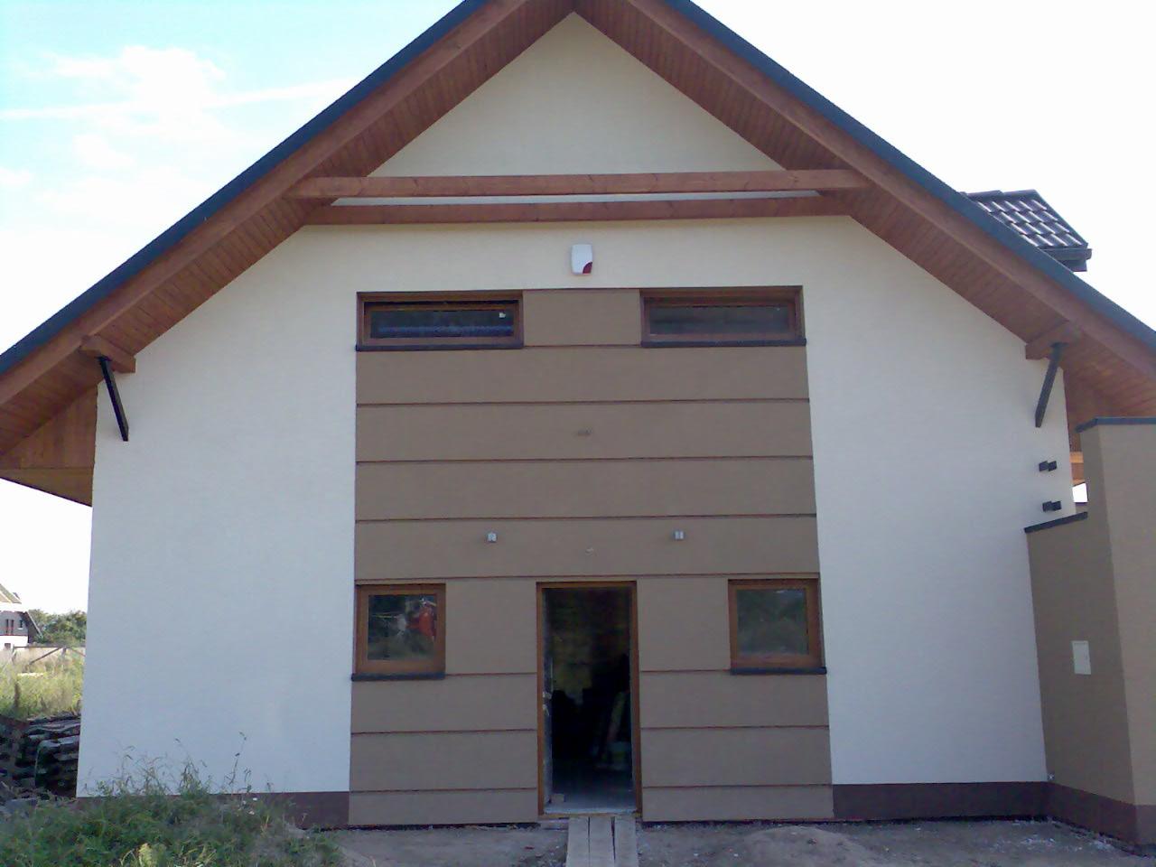 Fasada domu jednorodzinnego z drewnianymi oknami i elementami dekoracyjnymi. Jasne tynki kontrastują z brązowymi panelami.