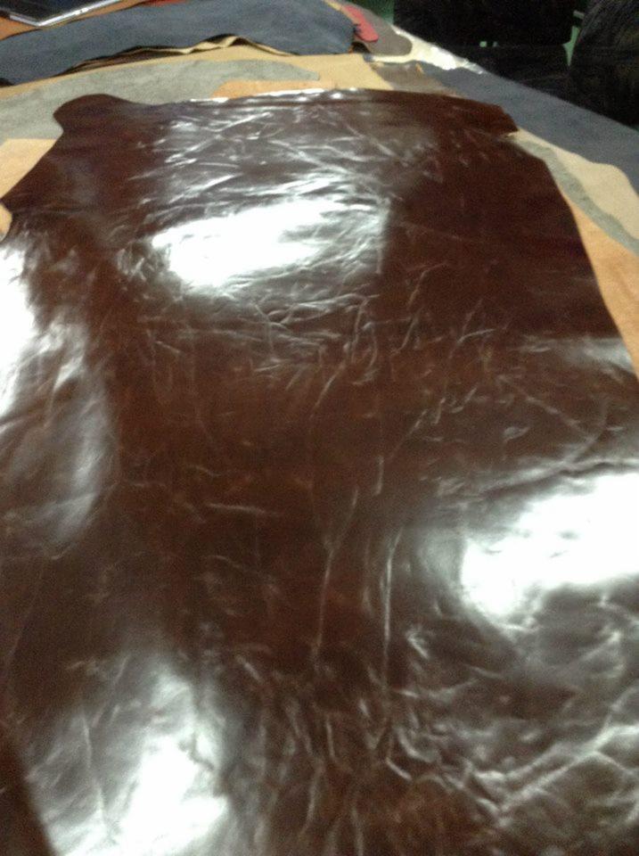 Błyszczący arkusz brązowej skóry naturalnej z delikatnymi rysami, leżący na stole w pracowni tapicerskiej. W tle widoczne fragmenty innych materiałów.