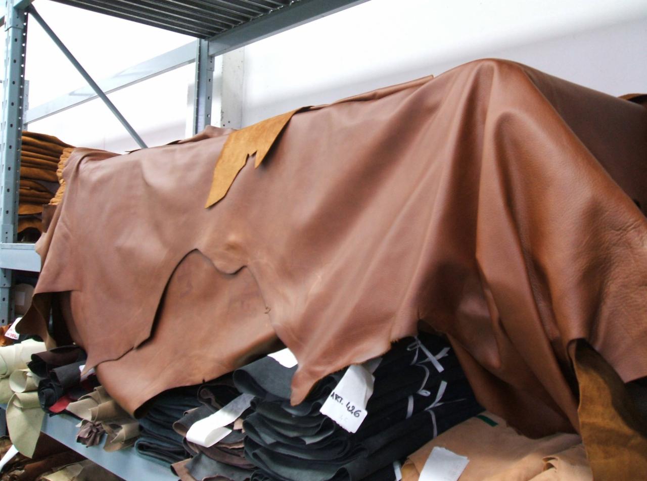 Składowane skóry naturalne w magazynie, różne kolory i faktury. Na półce widoczne etykiety z oznaczeniami artykułów. Oświetlenie magazynowe, metalowe regały.