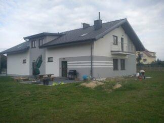 Dwukondygnacyjny dom po ociepleniu elewacji, z szarymi pasami, balkonem i ciemnym dachem, widoczny z trawnika w pochmurny dzień.