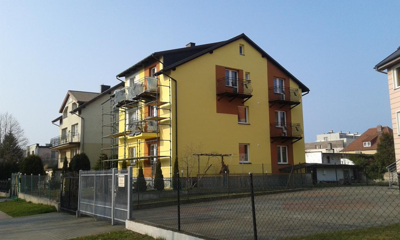 Żółta elewacja budynku z balkonami w trakcie prac, widoczne rusztowania. Częściowo odnowiona fasada, kontrastowe brązowe elementy. Słoneczny dzień.