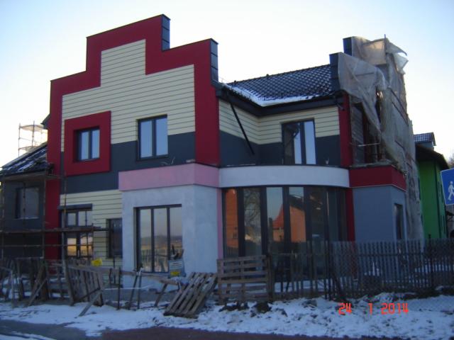 Wielobarwna elewacja domu jednorodzinnego w trakcie prac wykończeniowych, z widocznymi rusztowaniami i materiałami budowlanymi na pierwszym planie.
