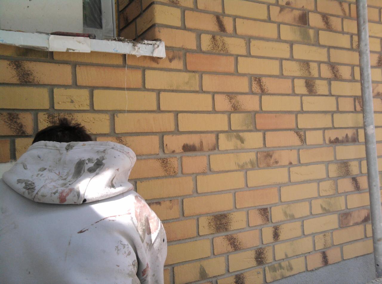 Mężczyzna w ubrudzonej farbą bluzie stoi tyłem przy murze z żółtej cegły z ciemnymi przebarwieniami, prawdopodobnie podczas renowacji elewacji.