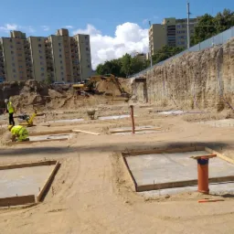 Przygotowanie terenu pod budowę: wykopy pod fundamenty w piasku, widoczny ciężki sprzęt i pracownicy w odblaskowych kamizelkach, w tle budynki mieszkalne.