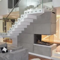 Nowoczesne schody z białymi stopniami i szklanymi balustradami, wbudowane w szarą, betonową konstrukcję z przeszklonym kominkiem, widok z salonu z szarą sofą.