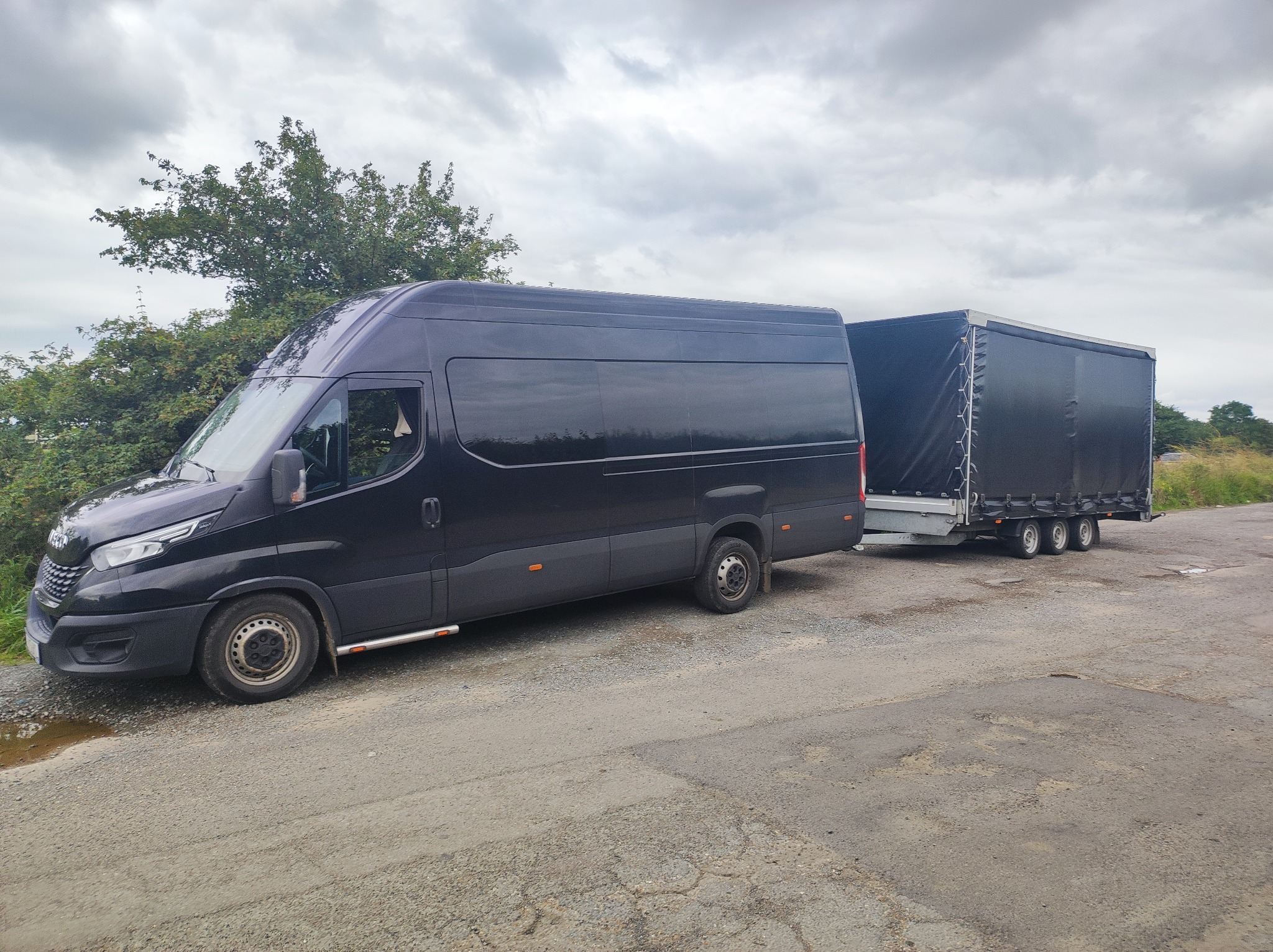 Czarny bus z przyczepą plandekową zaparkowany na poboczu drogi, widoczny fragment drzewa i pochmurne niebo w tle.