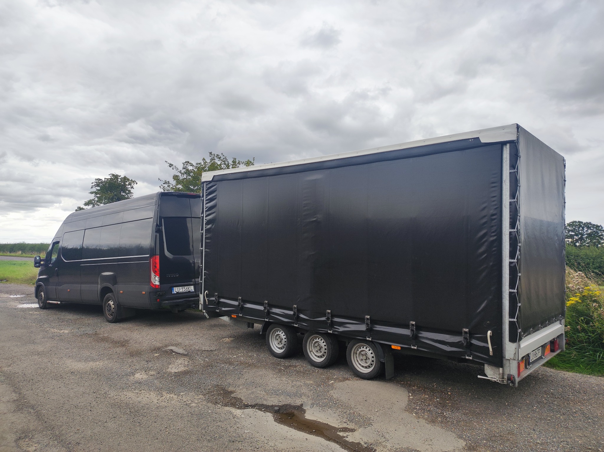 Czarny bus Iveco z otwartymi drzwiami połączony z trzyosiową przyczepą z czarną plandeką na szarym żwirowym podłożu i pochmurnym niebem w tle.