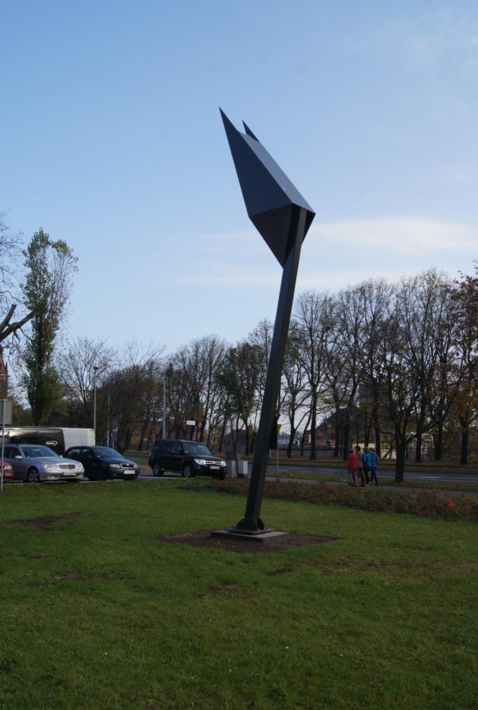 Metalowa wieża lęgowa dla jerzyków o geometrycznym kształcie, wznosząca się na trawiastym terenie miejskim z widocznymi samochodami i przechodniami w tle.