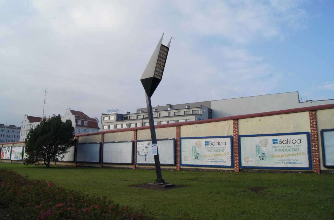 Wysoka, pochylona wieża lęgowa dla jerzyków w Gdańsku, ustawiona na trawniku, w tle budynki i reklamy okien Baltica.