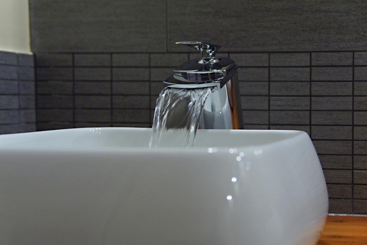 Biała, kwadratowa umywalka z chromowaną baterią wylewającą strumień wody, na tle ściany z szarych, prostokątnych płytek.